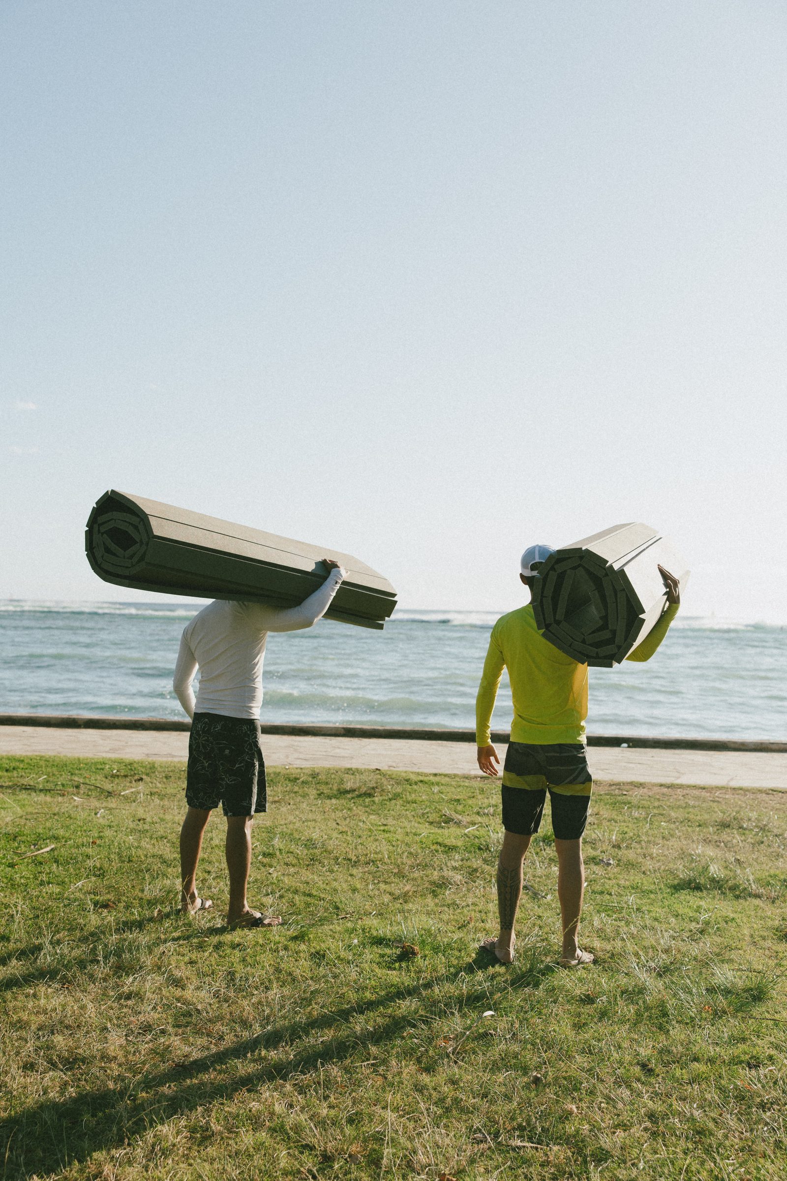 Two training partners carrying rolled mats across the grass to a beach session